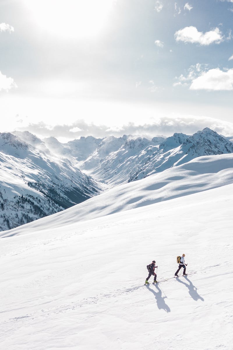 Home - english Two skiers climbing a sunlit snowy mountain slope in Ischgl, Austria, during winter.